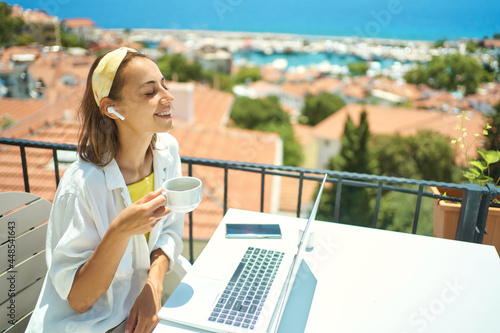 Pleased woman enjoying morning coffee while sitting on terrace with sea view on Turkish resort, using laptop computer for remote work, freelance concept