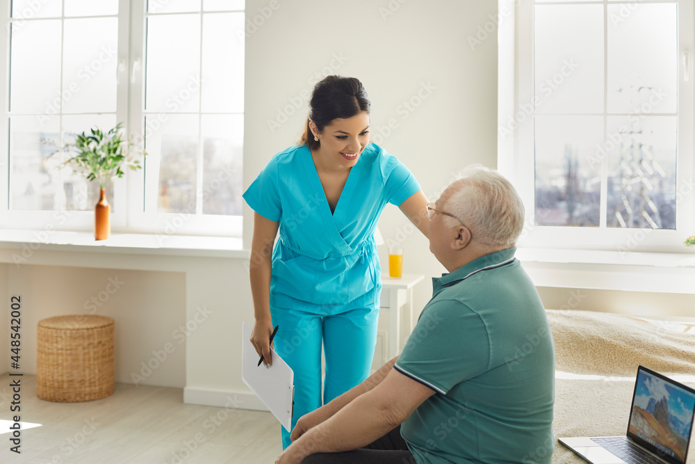 Fototapeta premium Woman doctor nurse asking for old patient well-being sitting on bed at hospital ward. Caregiver and aged man have friendly talk about health feeling. Morning medical check-up in nursing home
