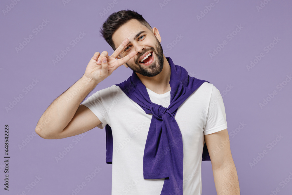 Attractive smiling happy young brunet man 20s wears white t-shirt purple shirt do hide eye behind victory sign isolated on pastel violet background studio portrait. People emotions lifestyle concept Attractive smiling happy young brunet man 20s wears white t-shirt purple shirt do hide eye behind victory sign isolated on pastel violet background studio portrait. People emotions lifestyle concept