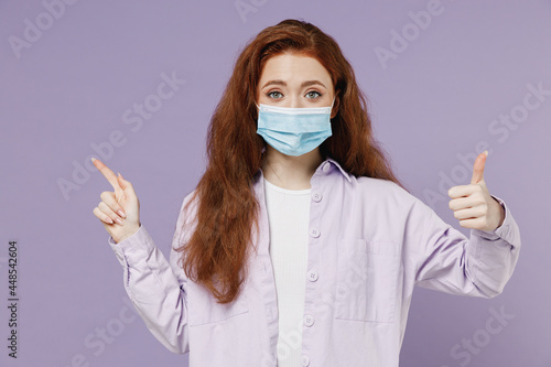 Young redhead woman 20 wear white T-shirt jacket sterile face mask ppe to safe from covid-19 flu point on workspace area copy space mock up isolate pastel purple color wall background studio portrait