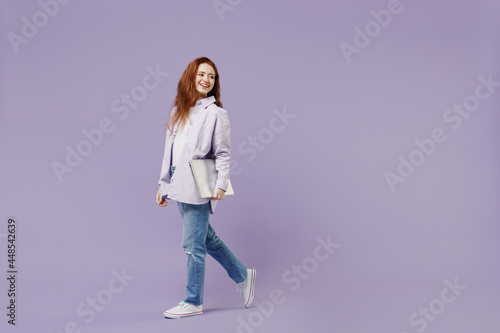 Full size body length fun young redhead curly woman 20 wear white T-shirt violet jacket walk go pace hold laptop pc computer under hand isolated on pastel purple color wall background studio portrait