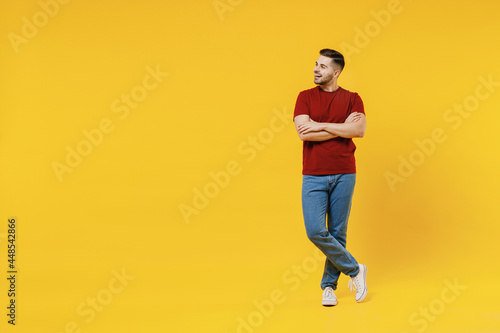 Full length smiling happy young man 20s wear red t-shirt casual clothes hold hands crossed folded look aside isolated on plain yellow color wall background studio portrait. People lifestyle concept.