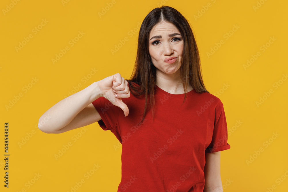 Sceptical dissatisfied discontent young brunette woman 20s wears basic red t-shirt showing thumb down dislike gesture isolated on yellow background studio portrait. People emotions lifestyle concept. Sceptical dissatisfied discontent young brunette woman 20s wears basic red t-shirt showing thumb down dislike gesture isolated on yellow background studio portrait. People emotions lifestyle concept.