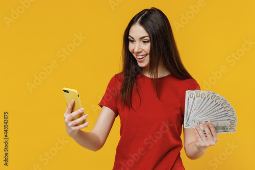 Obraz na plátně Satisfied smiling excited charming young brunette woman 20s wears basic red t-shirt hold use mobile cell phone and fan of cash money in dollar banknotes isolated on yellow background studio portrait