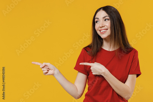 Smiling joyful young brunette woman 20s wears basic red t-shirt point look aside on workspace area copy space mock up isolated on yellow background studio portrait. People emotions lifestyle concept.