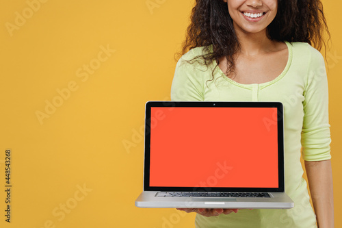 Close up jubilant cheery african american young brunette woman 20s wears green shirt hold use work on laptop pc computer with blank screen workspace area isolated on yellow background studio portrait. © ViDi Studio