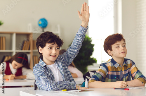 Canvas-taulu Smart schoolboy sits at a desk next to his classmate in class with his hand raised, wanting to give the right answer or participate
