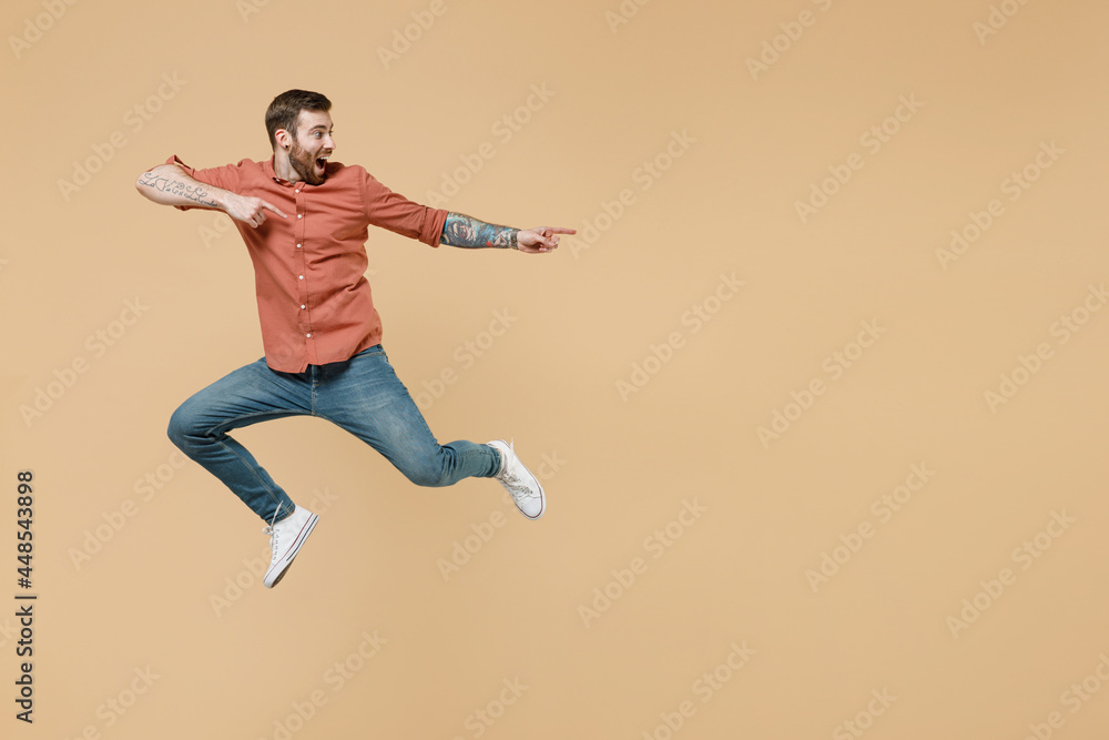 Full size body length tatooed young brunet man 20s short haircut wear apricot shirt jump pointing back behind on workspace area copy space mock up isolated on pastel orange background studio portrait