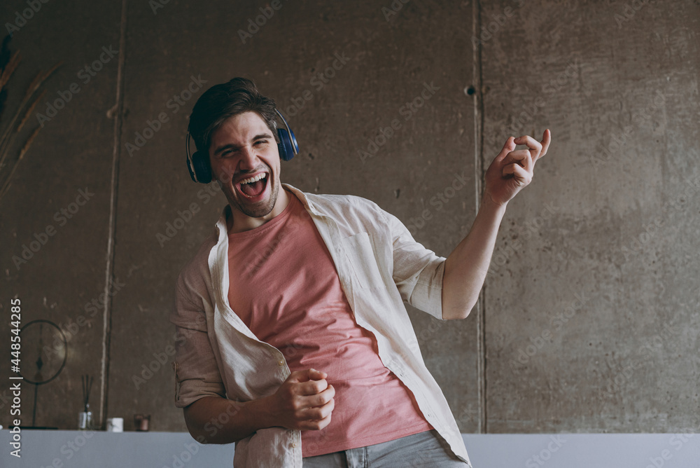 Young expressive singer fun joyful happy overjoyed man wear casual clothes beige shirt pink t-shirt sing headphones listen music play guitar rest indoors at home on weekends on brown wall background Young expressive singer fun joyful happy overjoyed man wear casual clothes beige shirt pink t-shirt sing headphones listen music play guitar rest indoors at home on weekends on brown wall background