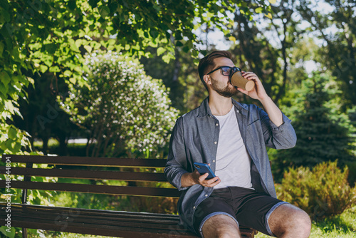 Young man in blue shirt shorts glasses sit on bench hold mobile phone takeaway cup drink coffee rest relax in spring green city park sunshine lawn outdoor on nature Urban lifestyle leisure concept photo