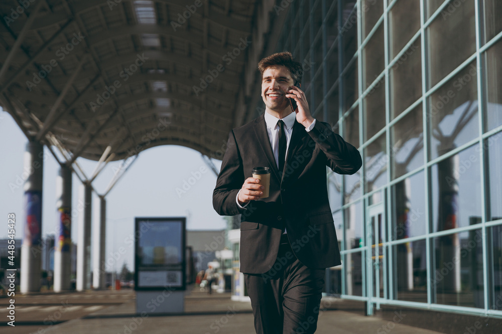 Young traveler businessman man wear black suit walk go outside at international airport terminal use mobile cell phone book taxi order hotel hold paper cup of coffee Air flight business trip concept. Young traveler businessman man wear black suit walk go outside at international airport terminal use mobile cell phone book taxi order hotel hold paper cup of coffee Air flight business trip concept.