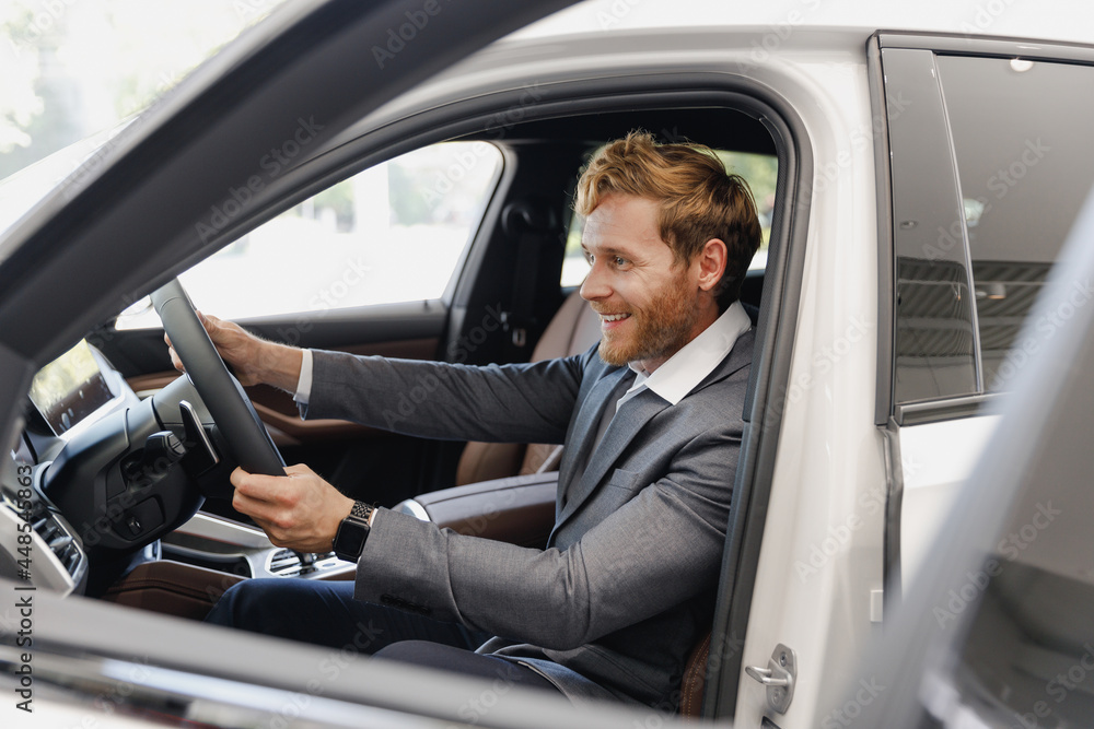 Side view man happy customer male buyer client in classic grey suit driving car hold wheel choose auto want buy new automobile in showroom vehicle salon dealership store motor show indoor Sale concept Side view man happy customer male buyer client in classic grey suit driving car hold wheel choose auto want buy new automobile in showroom vehicle salon dealership store motor show indoor Sale concept