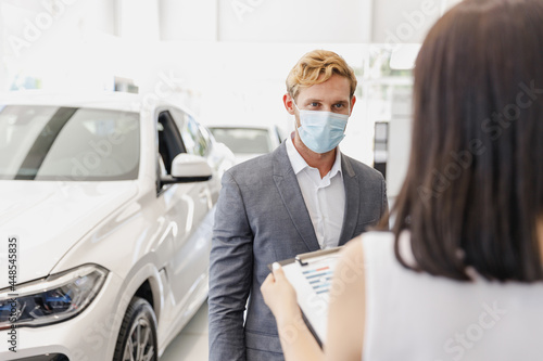 Man customer client buyer in classic suit pandemic mask choose auto want buy new car automobile consult with salesman read papers in showroom salon dealership store motor show indoor Sale concept.
