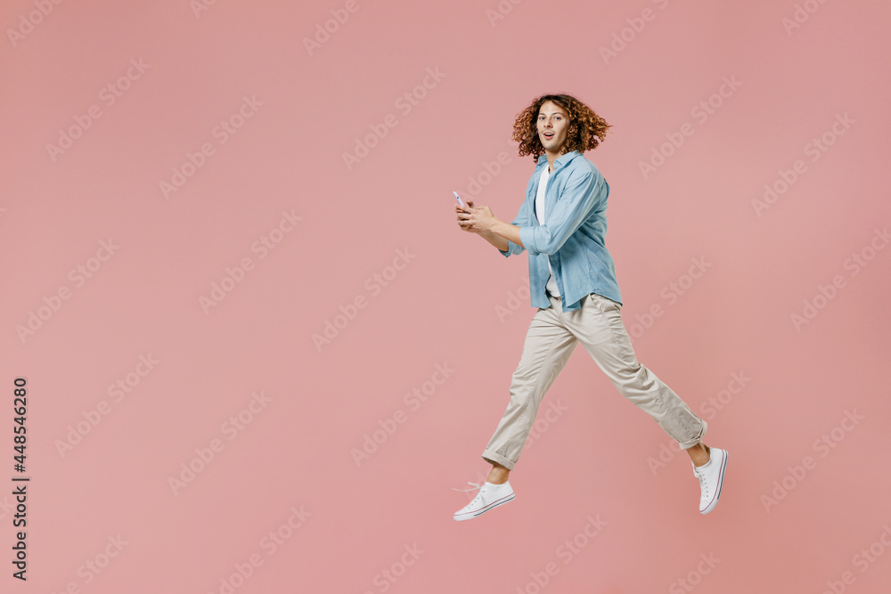Full length side view young excited happy man with long curly hair wear blue shirt white t-shirt using hold in hand mobile cell phone jump high isolated on pastel plain pink color background studio