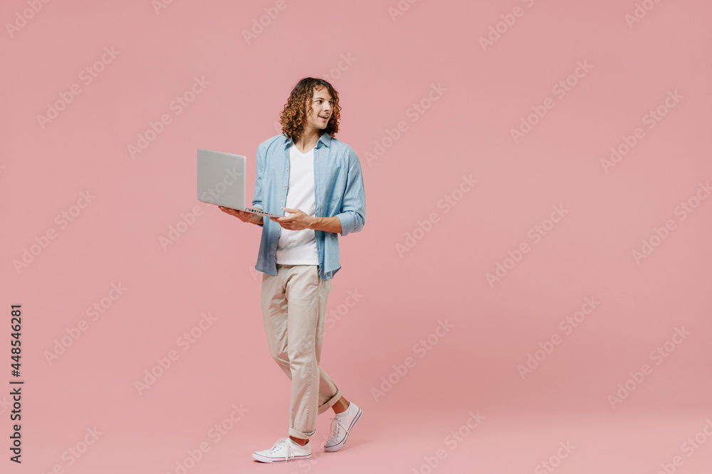 Fototapeta premium Full length young minded happy man with long curly hair in blue shirt white t-shirt hold use work on laptop pc computer look aside isolated on pastel plain pink color wall background studio portrait
