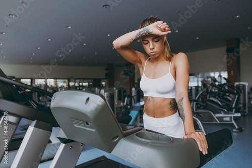 Young sweating strong sporty athletic sportswoman woman in white sportswear warm up training run on treadmill put hand on forehead drink water in gym indoor Workout sport motivation lifestyle concept photo