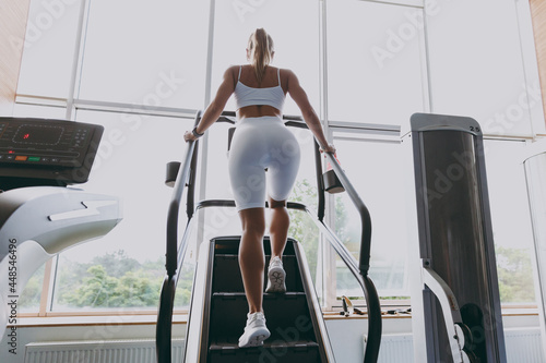 Back view full size young strong sporty athletic sportswoman woman in white sportswear warm up training running on a treadmill climber stairs machine in gym indoors. Workout sport motivation concept.