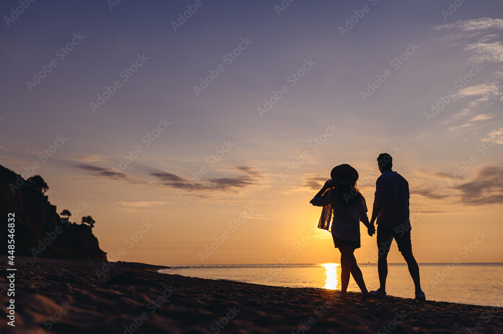 Full size back view silhouette young couple two friends family man woman in casual clothes hold hands walk stroll together at sunrise over sea beach ocean outdoor exotic seaside in summer day evening. Full size back view silhouette young couple two friends family man woman in casual clothes hold hands walk stroll together at sunrise over sea beach ocean outdoor exotic seaside in summer day evening.
