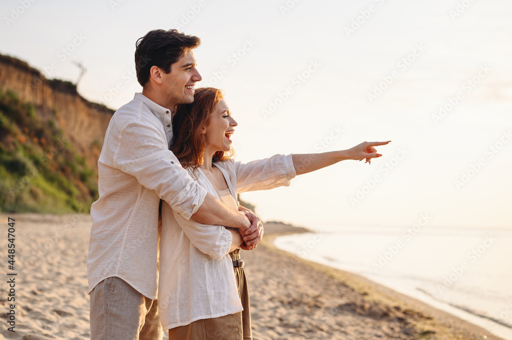Profile happy smiling young couple two friends family man woman in white clothes hug rest relax together point finger aside at sunrise over sea beach ocean outside seaside in summer day sunset evening