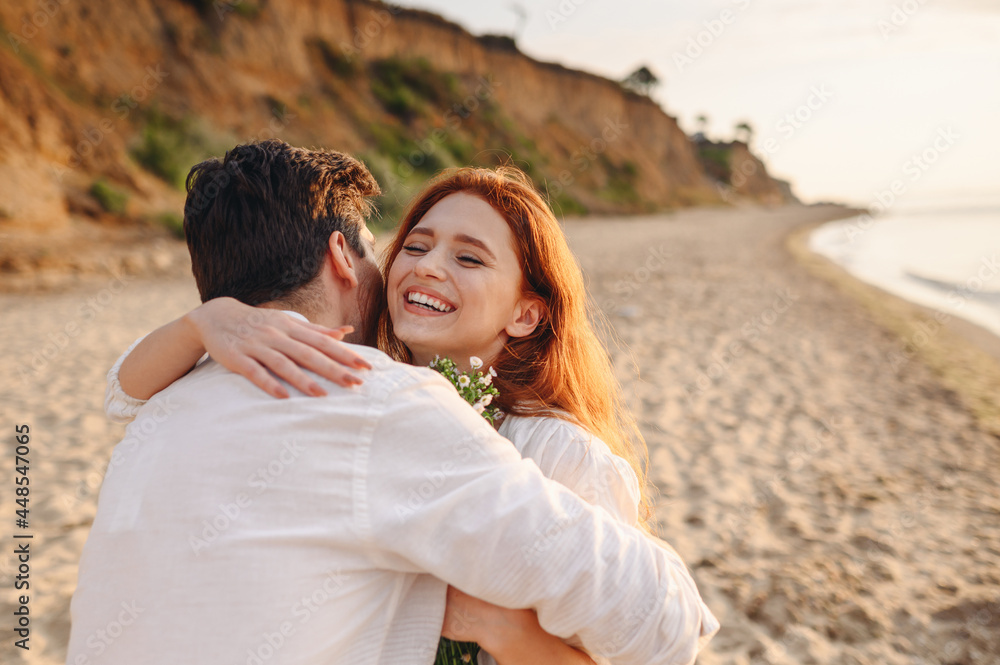 Excited young lovely couple two family man woman in white clothes hug each other hold flowers enjoy together at sunrise over sea ocean sand beach outdoor exotic seaside in summer day sunset evening.