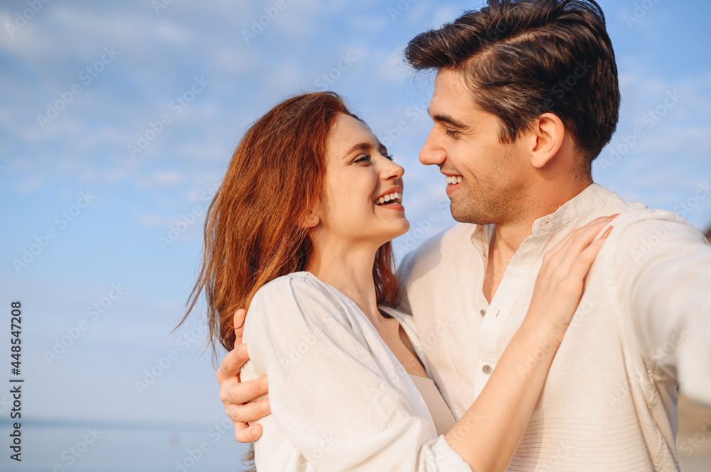 Close up enamored young couple two family man woman in white clothes hug look to each other do selfie shot pov on mobile phone at sunrise over sea beach outdoor seaside in summer day sunset evening. Close up enamored young couple two family man woman in white clothes hug look to each other do selfie shot pov on mobile phone at sunrise over sea beach outdoor seaside in summer day sunset evening.