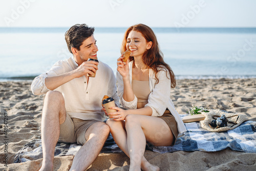 Young happy fun couple family two man woman in white clothes hug sitting sand plaid have picnic eat croissant drink coffee rest together at sunrise over sea beach ocean outdoor seaside in summer day