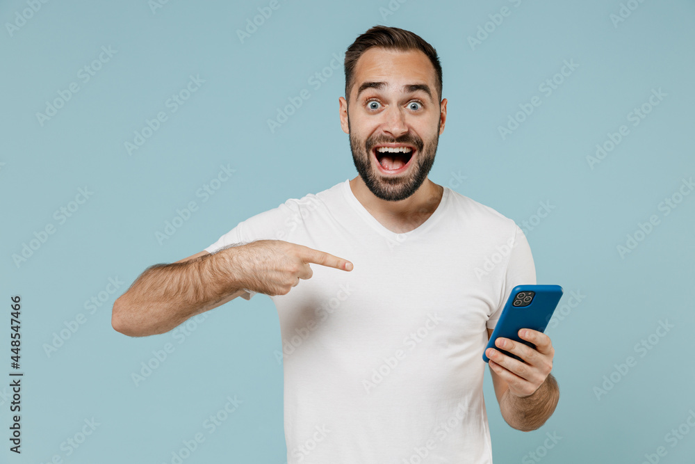Young surprised happy man 20s wear white casual blank print design t-shirt point index finger on mobile cell phone chatting online isolated on plain pastel light blue color background studio portrait Young surprised happy man 20s wear white casual blank print design t-shirt point index finger on mobile cell phone chatting online isolated on plain pastel light blue color background studio portrait