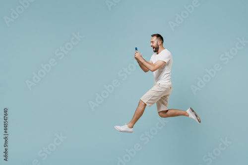 Full length side view young overjoyed happy man 20s wearing casual white t-shirt jump high using mobile cell phone chatting online isolated on plain pastel light blue color background studio portrait