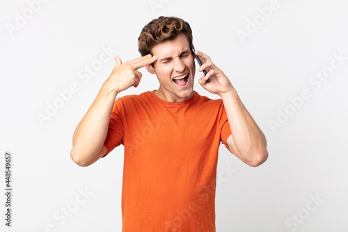 young handsome man looking unhappy and stressed, suicide gesture making gun sign and talking with a smart phone photo