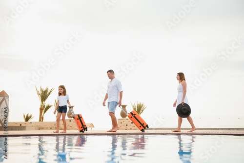 young family with suitcases walking to hotel building with beautiful swimming pool. traveling and relaxing time concept.