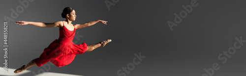 Side view of brunette ballerina in red dress jumping on grey background with sunlight, banner
