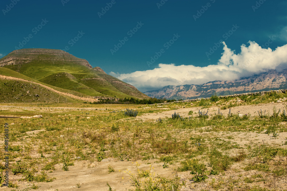 Toned mountain landscape view. Summer cloudy day in valley. Panorama with high hills, light brown earth, green grass and background of blue sky with white clouds.