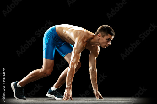 Side view of young professional male athlete, runner preparing to start isolated on dark studio background. Muscular, sportive man. Concept of sport, healthy lifestyle