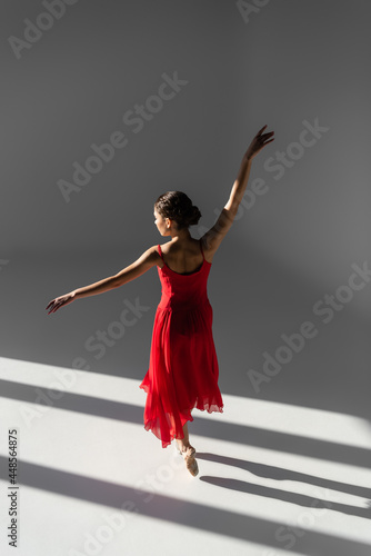 Ballerina in pointe shoes and red dress standing on grey background with sunlight