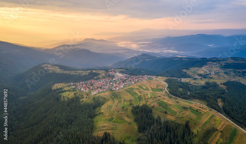 Aerial view of picturesque green mountain slopes and a small village nestled in them in golden hour at sunset, the Rhodopi Mountains in Bulgaria.