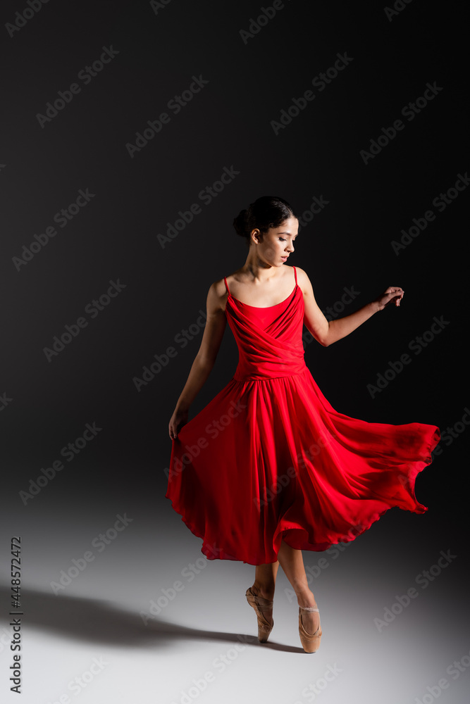 Naklejka premium Young brunette ballerina in red dress dancing on black background
