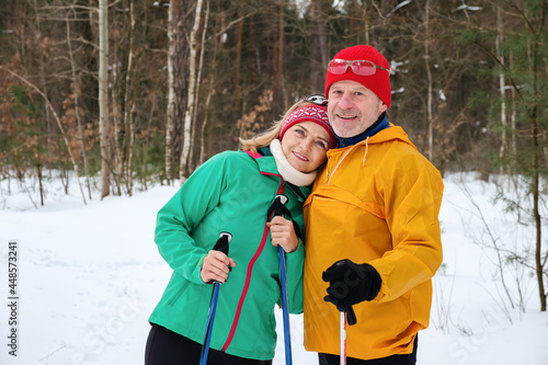 Smiling senior couple walking with nordic walking poles in snowy winter forest. Elderly wife and husband doing healthy exercise outdoors. Active lifestyle after retirement concept.