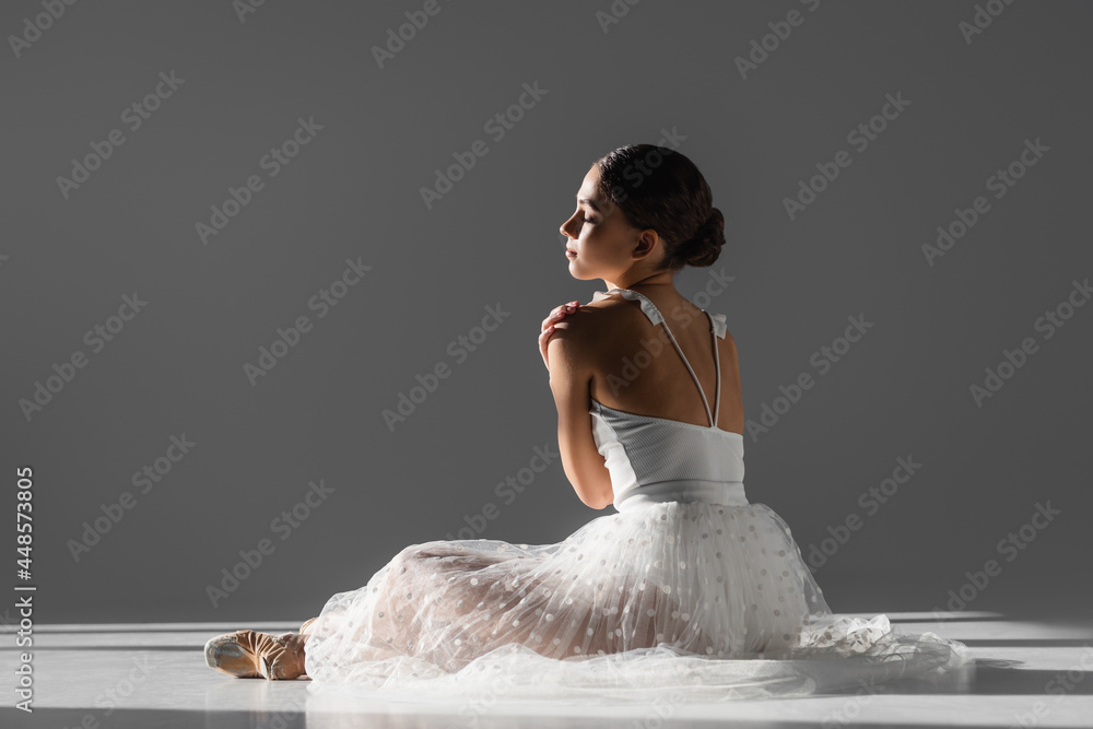 Naklejka premium Side view of young ballerina touching shoulder on grey background with sunlight