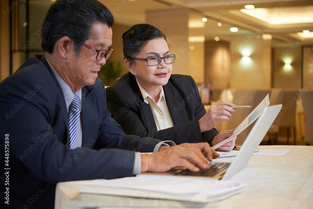 Senior businesswoman pointing at sales chart when talking to her colleague at meeting Senior businesswoman pointing at sales chart when talking to her colleague at meeting
