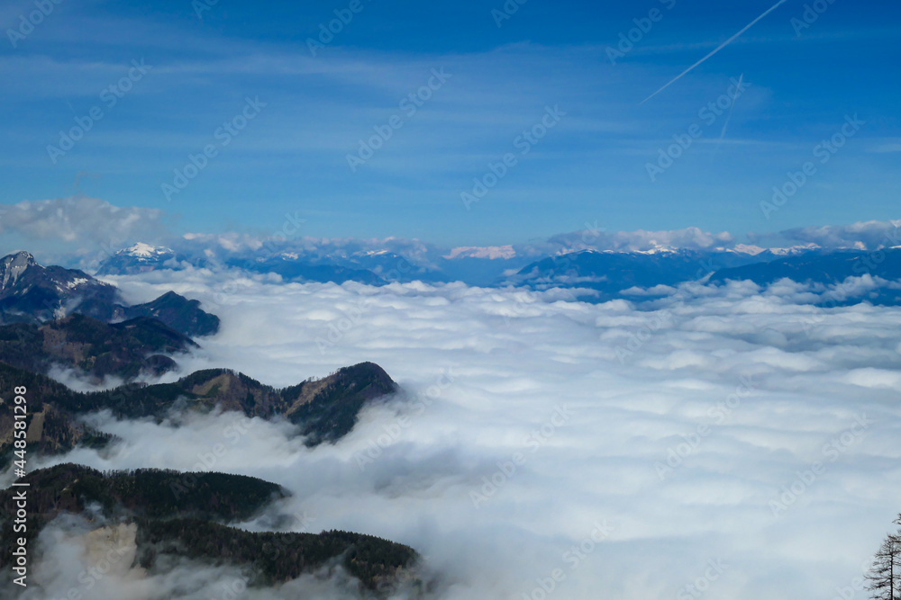 A panoramic view from the top of Alpine peak in Austria. The whole area is shrouded in thick clouds. A few peaks popping out from the clouds. High mountain chains in the back. Carpet from the clouds