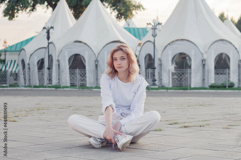A beautiful woman in white clothes is sitting on a concrete tile on the street
