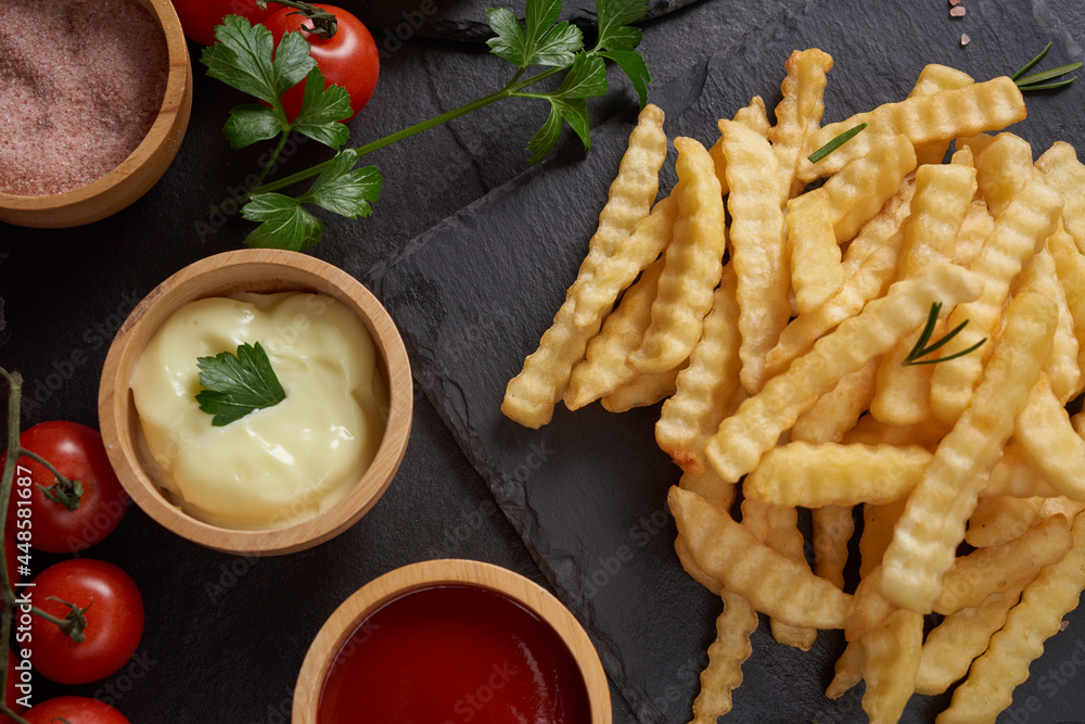 Homemade baked potato fries with mayonnaise, tomato sauce and rosemary on paper over black stone background. . tasty french fries, unhealthy food.