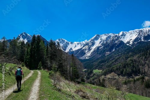 A woman trekking with a backpack on a hiking trail with the view on Baeren Valley in Austrian Alps. The highest peaks in the chain are snow-capped. A few trees on the slopes. Sunny day. Wanderlust