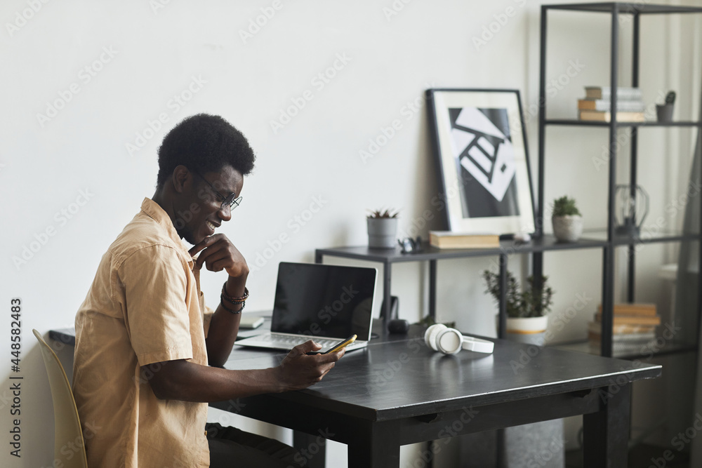 African happy man sitting at the table with laptop at office reading a message on mobile phone and smiling
