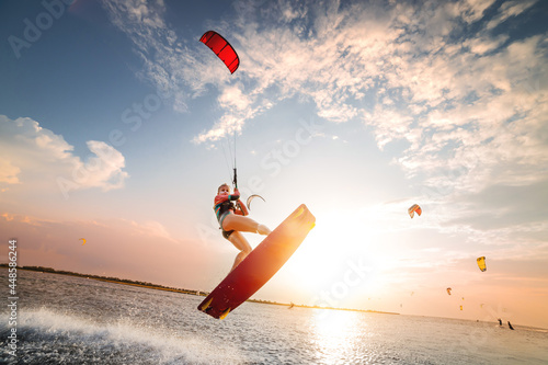 Sports shot of a young caucasian woman in a wetsuit doing a trick in the air against the backdrop of a sunset in the sea. Kitesurring girl athlete flies on a kite with a board