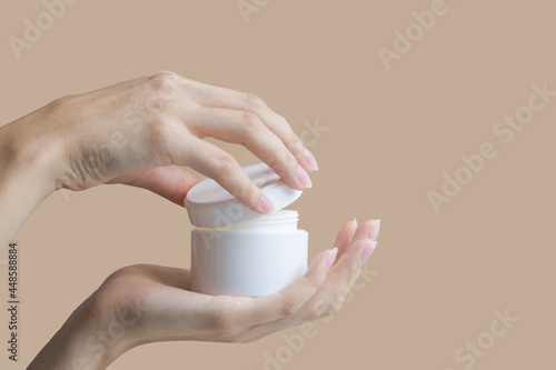 Women's hands open mock-up of white cosmetic package. Two hands hold round jar of body cream on beige background. Concept of beauty industry. photo