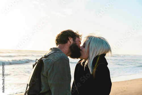 Romantic happy couple in love kissing on shoreline at the wild beach - Boyfriend and girlfriend wearing cool clothing and vintage backpack standing in front of the sea during the sunset