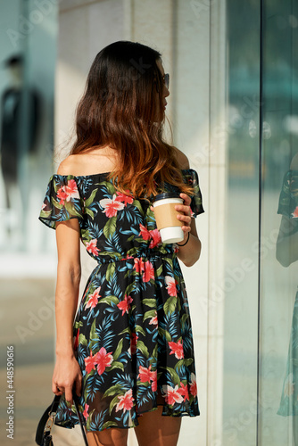 Pretty young woman in flower dress walking outdoors with cup of coffee and looking at show windows photo