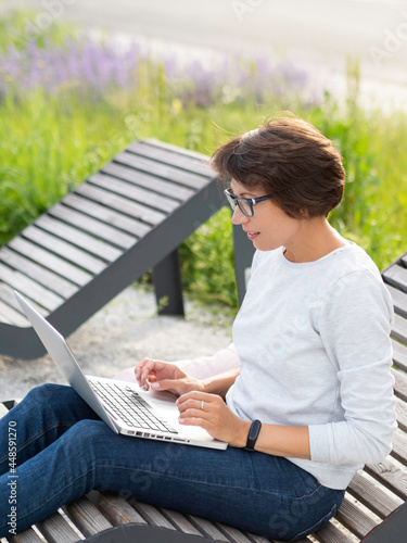 Woman sits with laptop on urban park bench. Freelancer at work. Student learns remotely from outdoors. Modern lifestyle.
