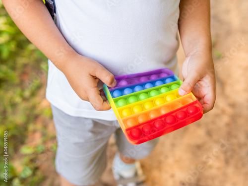 Little boy plays square rubber fidget toy Pop It. Rainbow-colored anti stress and tactile toy for all ages. Outdoor recreation with toy for fine motor skills improvement.